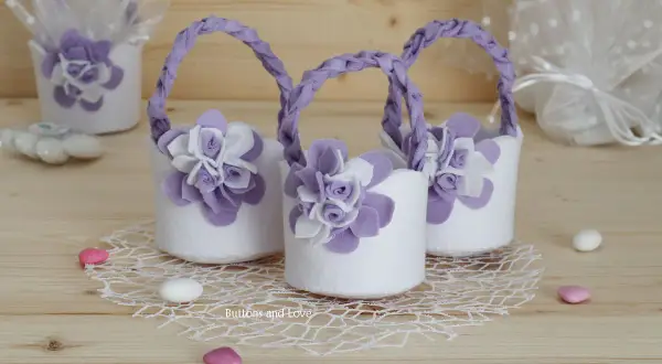 Piccoli cestini porta bomboniere fai da te in feltro bianco con fiori lilla e manici intrecciati, perfetti per un evento speciale.