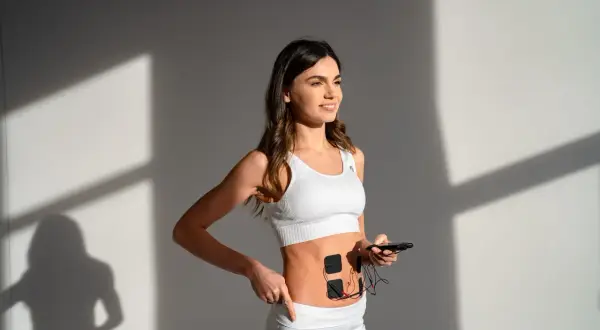 Elin w białym sportowym topie i legginsach, z elektrodami na brzuchu, trzyma smartfon.