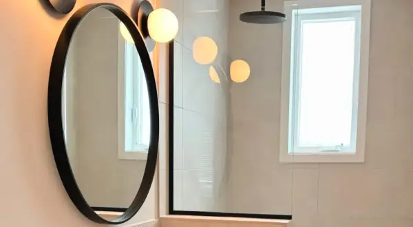 Un élégant miroir au-dessus du lavabo reflète des lumières douces. La salle de bain moderne comprend une douche vitrée et une fenêtre lumineuse.