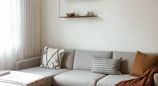 Gemütliches Wohnzimmer mit grauem Ecksofa, Kissen und Decken in warmen Erdtönen. Die schlichte Einrichtung könnte die **Farbe des Jahres 2026** widerspiegeln.