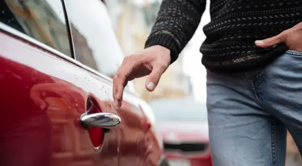 Mano tocando la manija de un vehículo rojo, con un suéter y jeans. Parece que el conductor está a punto de entrar.
