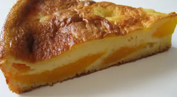 Tranche de clafoutis aux pêches, dorée et moelleuse, avec des morceaux de fruits juteux.