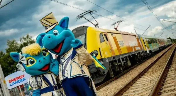 Dwa niebieskie smoki w strojach pozują przy żółtym pociągu. To specjalny pociąg do Energylandii, który zabierze Cię na niezapomnianą przygodę!