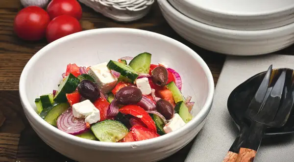 Frischer griechischer Salat mit Tomaten, Gurken, roten Zwiebeln, Oliven und Feta in einer weißen Schüssel.