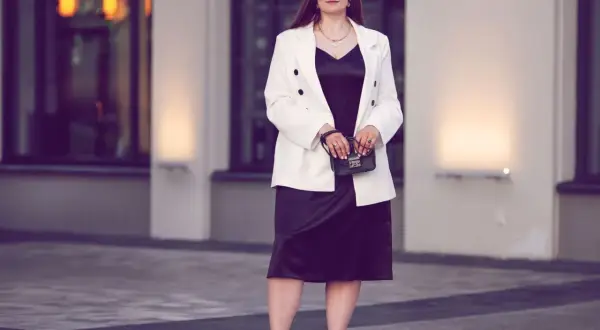 Eine Frau in einem schwarzen Kleid und weißem Blazer, die zeigt, was man zum Musical anzieht. Sie trägt weiße Sneaker und eine kleine Handtasche.