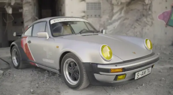 Srebrny Porsche 911 z żółtymi reflektorami, w stylu cyberpunk, stoi na tle zniszczonej ściany z graffiti.