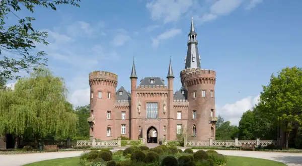 Schloss Moyland, ein imposantes Backsteinschloss mit Türmen und einem hohen Spitzdach, thront über einer gepflegten Gartenanlage mit einem runden Blumenbeet.