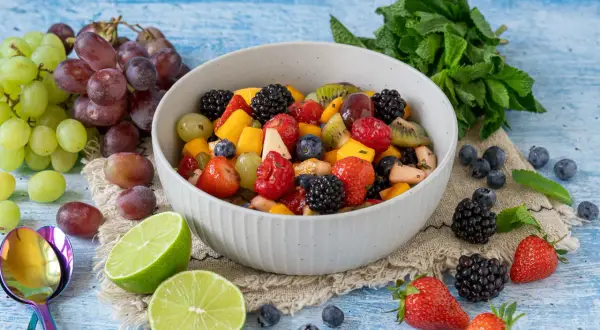 Ein bunter, sommerlicher Obstsalat mit Beeren, Trauben und Mango, garniert mit Minze und Limettenspalten.