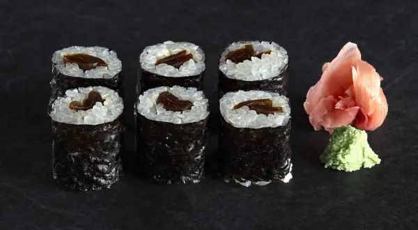 Sześć kawałków sushi z ryżem i ciemnym nadzieniem, obok różowy imbir i zielone wasabi.