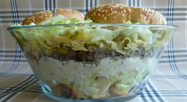 Warstwowa sałatka big mac przepis: mięso, ser, sałata, ogórek i sos w szklanej misie, zwieńczona bułkami z sezamem.