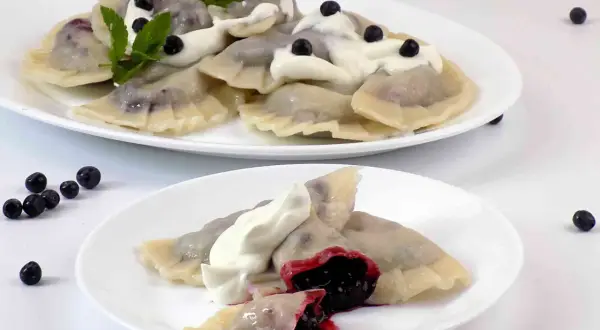 Pyszne pierogi z jagodami, polane śmietaną i ozdobione miętą. Na talerzu widać też kilka świeżych jagód.