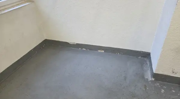 Ecke eines Raumes mit grauem Boden und weißer Wand. Am Sockel ist ein dunkler Streifen zu sehen, auf dem sich schimmel zieht von unten nach oben.