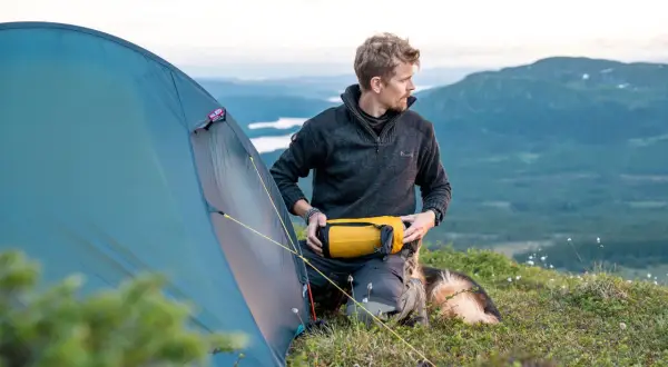 Man med hund förbereder tältet för en vandring i Norrland. Vacker fjällvy i bakgrunden.
