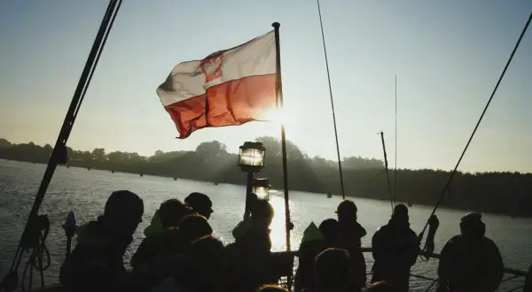 Słońce przebija się przez biało-czerwoną bandera polską na statku. Grupa ludzi obserwuje zachód słońca nad wodą.