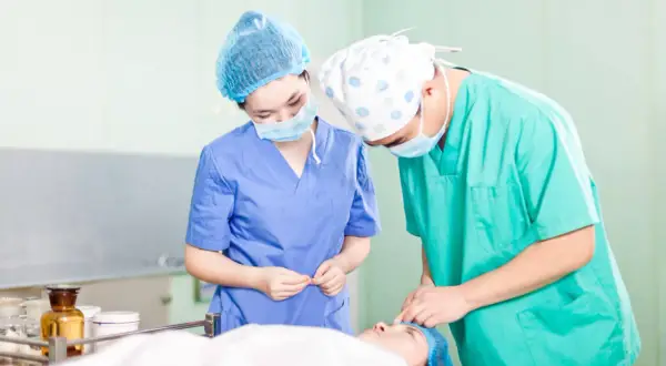 Deux chirurgiens masqués et coiffés préparent une intervention. L'un tient des instruments, l'autre s'occupe du patient. Ils se demandent "que veut dire DAE" dans ce contexte médical.