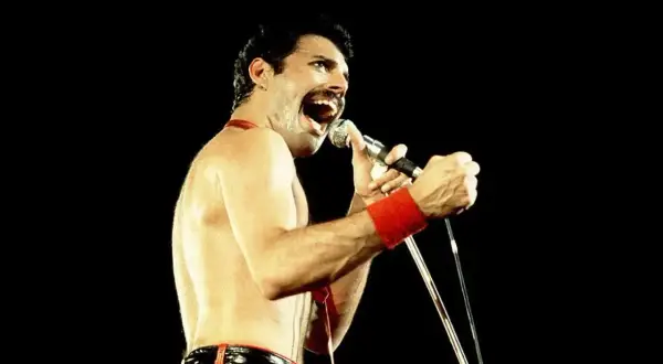 Freddie Mercury w czerwonych spodniach i opasce na nadgarstku śpiewa do mikrofonu. Nawet jako dorosły, jego energia przypominała dzikie dziecięce zabawy.