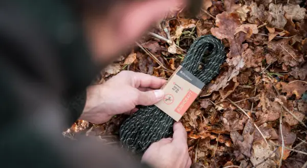 Manos sostienen un rollo de cordón paracord verde con puntos blancos, sobre un lecho de hojas secas.