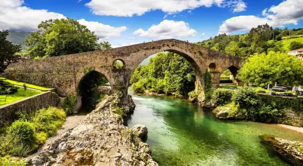 Majestuoso puente romano asturias sobre un río de aguas cristalinas, rodeado de exuberante vegetación y un cielo azul con nubes blancas.