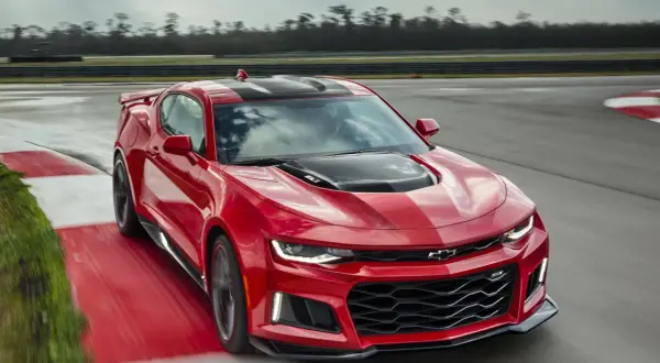 Czerwone Camaro ZL1 z czarnymi pasami na masce i dachu pędzi po torze wyścigowym, zostawiając za sobą smugę.