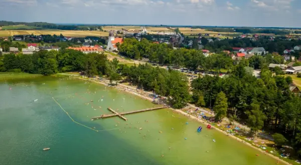 Tłum ludzi wypoczywa nad jeziorem wielewskim. Woda jest zielona, a na brzegu widać plażę i las. W oddali majaczy kościół.