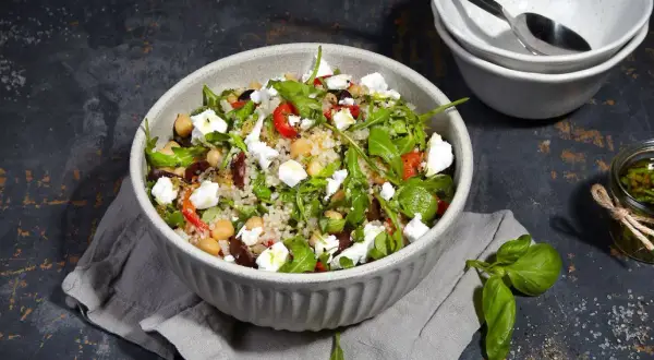 Bunter Quinoa Salat mit Kichererbsen, Feta, Oliven und Rucola, serviert in einer Schüssel. Daneben ein Glas mit Kräuter Dressing.