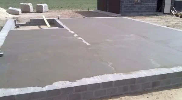 Świeżo wylany chudy beton na fundamencie domu. W tle widać materiały budowlane i fragment budynku.