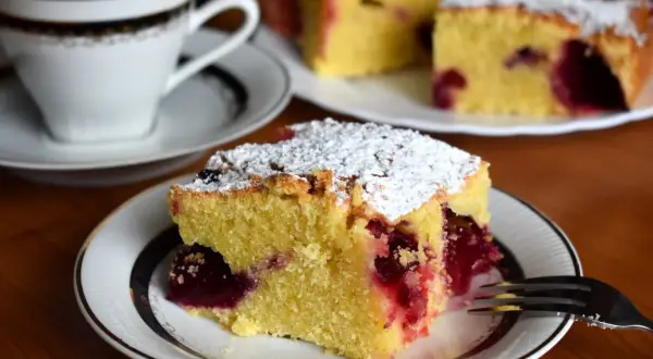 Szybkie ciasto bezglutenowe z wiśniami, posypane cukrem pudrem, na talerzyku obok filiżanki kawy. Idealne na deser.