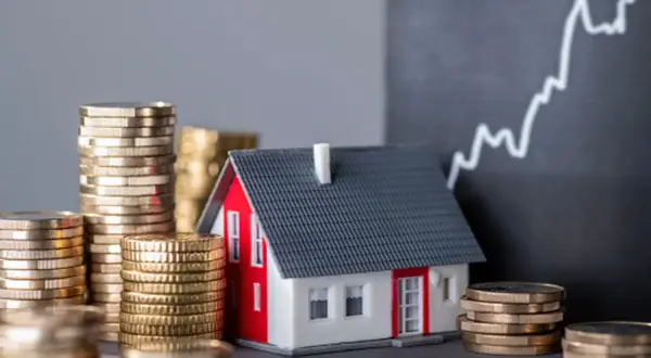 Casa modelo rodeada de pilas de monedas, con un gráfico ascendente al fondo, sugiriendo un estudio de solvencia y el valor de la inversión inmobiliaria.
