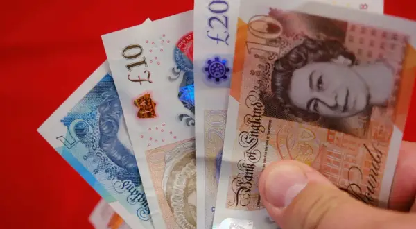 Dłoń trzyma banknoty: 5, 10 i 20 funtów. Różne nominały funtów widać na czerwonym tle.