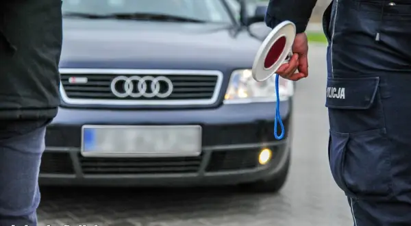 Policjant z lizakiem zatrzymuje auto. Widać jego mundur i fragment ciemnego Audi. Jakie światła podczas deszczu, gdy policja kontroluje ruch.