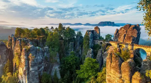 Felsformationen und eine Basteibrücke in der Böhmischen Schweiz, umgeben von Nebel und Bäumen, unter einem blauen Himmel.
