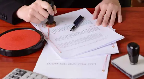 Dłoń z pieczęcią nad dokumentem, na którym widnieje napis "LAST WILL AND TESTAMENT". Obok poduszka z tuszem i długopis. To akt notarialny o poddaniu się egzekucji.