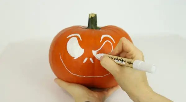 Ręce malują białą farbą wesołą buźkę na pomarańczowej dyni. Powstaje Halloweenowa dekoracja.