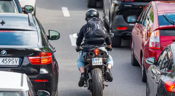 Motocyklista w kasku i plecaku przeciska się między samochodami w korku. Widać znaki motocyklistów na tablicy rejestracyjnej.
