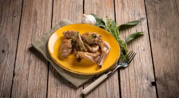 Sabroso conejo al ajillo servido en plato amarillo, acompañado de hierbas frescas y un tenedor, sobre una mesa de madera rústica.