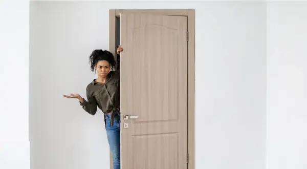 Mujer asomándose por una puerta de madera clara, con expresión de duda. Los colores de puertas son neutros y elegantes.
