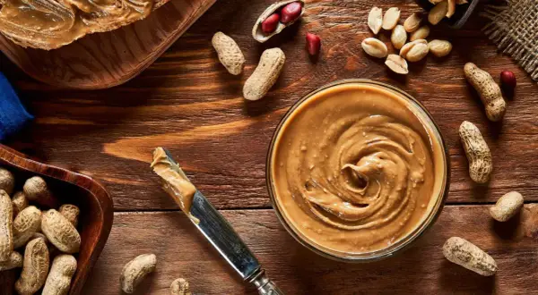Cremige Erdnussbutter in einer Schüssel, daneben Erdnüsse und ein Messer. Erdnussbutter gesund und lecker!