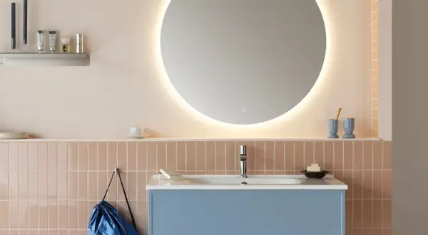 Ett modernt badrum med en stor, upplyst rund spegel som ger en känsla av rymd. Att inreda med speglar är smart! Blått kommod och rosa kakel skapar en mjuk atmosfär.