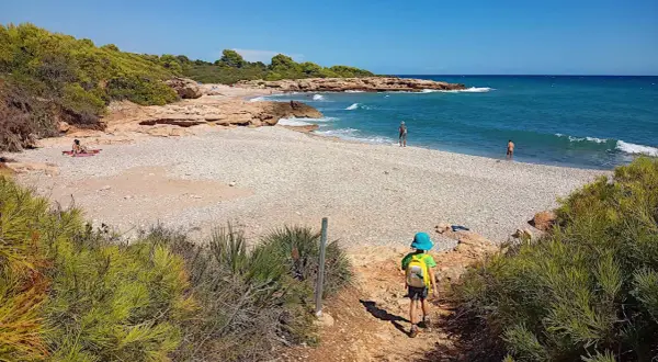 Niño con mochila azul desciende por sendero hacia la playa de rueta, con rocas y mar turquesa.