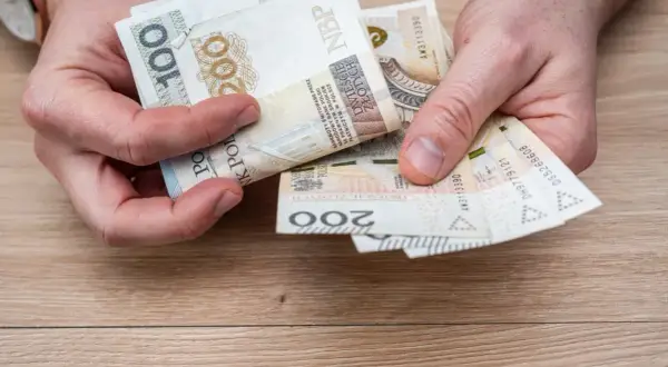 Dłonie trzymające polskie banknoty: 100 i 200 złotych. Widać fragment zegarka na ręce.