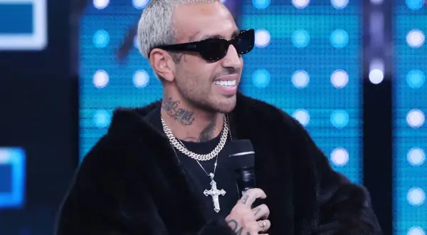 Uomo con capelli biondi platino, occhiali da sole neri e pelliccia nera. Indossa una collana d'oro con croce. Ti voglio luche!