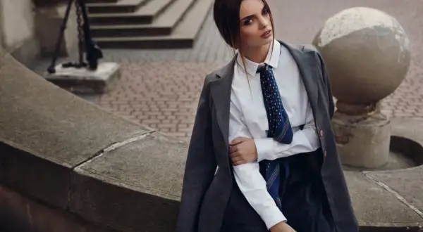Elegancka kobieta w białej bluzce z krawatką i szarej marynarce, siedząca na kamiennej ławce.