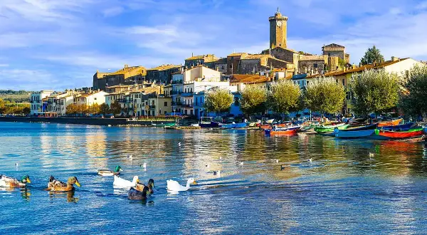 Anatre nuotano in un lago sereno, con un borgo pittoresco e barche colorate che si specchiano sull'acqua. Un'immagine da cartolina dei laghi italiani.