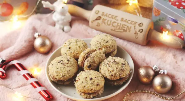 Leckere Nutella Kekse, umhüllt von gehackten Nüssen, liegen auf einem Teller. Ein Nudelholz mit der Aufschrift "Backe, backe Kuchen" und Weihnachtsdeko sorgen für festliche Stimmung.