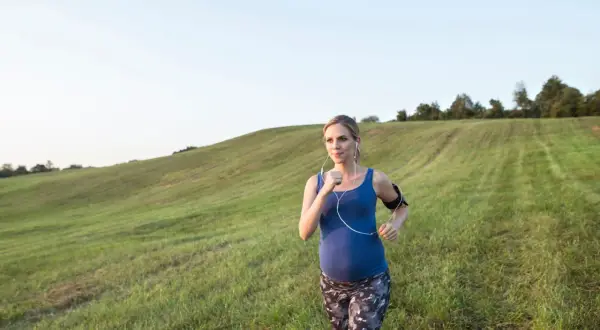 Donna incinta corre in un prato verde, ascoltando musica con le cuffie. Un'immagine di benessere e forza durante la gravidanza.