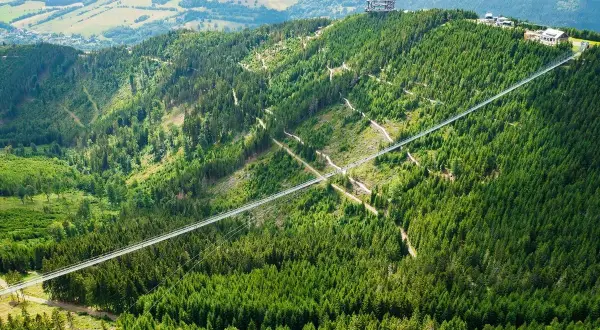Najdłuższy most w Czechach, Sky Bridge 721, przecina zieloną dolinę pełną drzew. Na szczycie góry widać wieżę widokową.