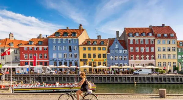 En kvinna cyklar längs Nyhavn i Köpenhamn, ett soligt exempel på danmark klimat. Färgglada hus och kanalbåtar skapar en livlig atmosfär.