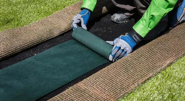 Manos enguantadas desenrollan césped artificial verde sobre una base negra. Se prepara para poner césped artificial sin pegar.