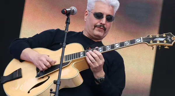 Un cantautore napoletano famoso con capelli bianchi e occhiali da sole suona una chitarra jazz.