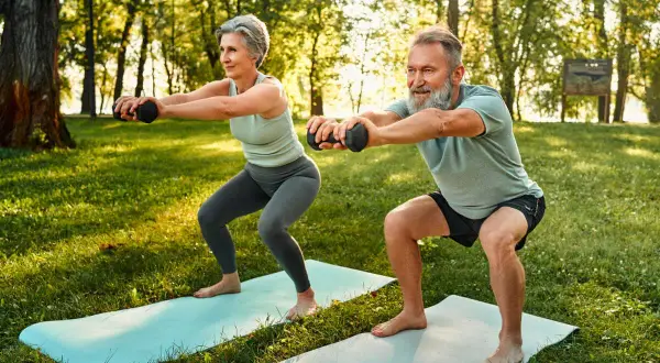 Une femme sportive de 50 ans et un homme font des squats avec des haltères dans un parc ensoleillé.